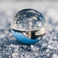 Color photo of an upside-down house in a glass ball with blue sky background. Top half of the ball looks like water and dark clouds.