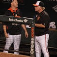 Then-Orioles manager Buck Showalter (right) stands in the Orioles dugout at Camden Yards, talking to another player. Showalter is wearing a uniform with white pants with an organze strpe, a black pullover windbreaker with the modern cartoon version of the Orioles’ mascot, and a cap in orange, black and white.