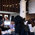 Photo of students speaking with employers and recruiters at a STEM employers event. People are gathered around tall tables indoors. There are lights strung from the ceiling.