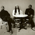Three men who are black sit at a white coffee table that forms a circle. The two men on the outside have short hair while the one in the middle has long dreads. This picture of the Broun Fellinis has a sepia tone.