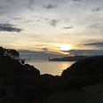 Sunset near the edge of a cliff, strewn with foliage, almost black in the fading light. The calm waters of the San Francisco Bay loom beyond, and the Golden Gate Bridge silhouetted in the distance, framed by a looming cloud bank, nigh obscuring the last bright remnants of the hot, white sun ball.