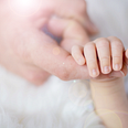 A brightly lit photo of an adult holding an infant’s hand with their pointer finger.
