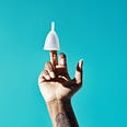 A woman’s hand and tattooed wrist with middle finger extended. On the tip of the middle finger is a menstrual cup.