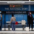 Passengers wearing face masks wait for their bus in front of a national debt display on Pennsylvania Ave. NW in Washington on Monday, May 18, 2020.