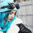 Woman with long electric blue hair in a black leather jacket and cyberpunk spike goggles over a blue beanie looking away from the camera