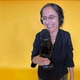 Standing against a yellow background, a laughing middle aged, white woman in pink glasses, black button down shirt, and headphones holds a large microphone