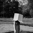 A woman stands in a field with a cardboard box on her head — trying to block the world out