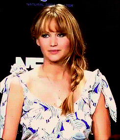 Jennifer Lawrence in a flowy blue & white dress exaggeratedly saying okay & giving a thumbs up. Her hair is in a side braid