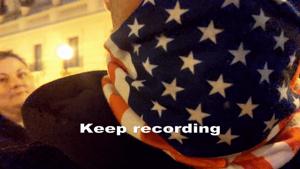 A close-up shot of a man in an American Flag mask with text that repeats “Keep recording! You ain’t gonna do nothing!”