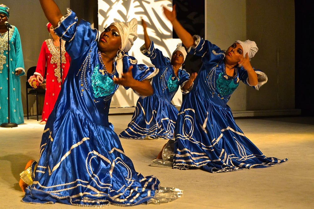 Three women in blue dresses stand in a slight lunge lifting one arm up