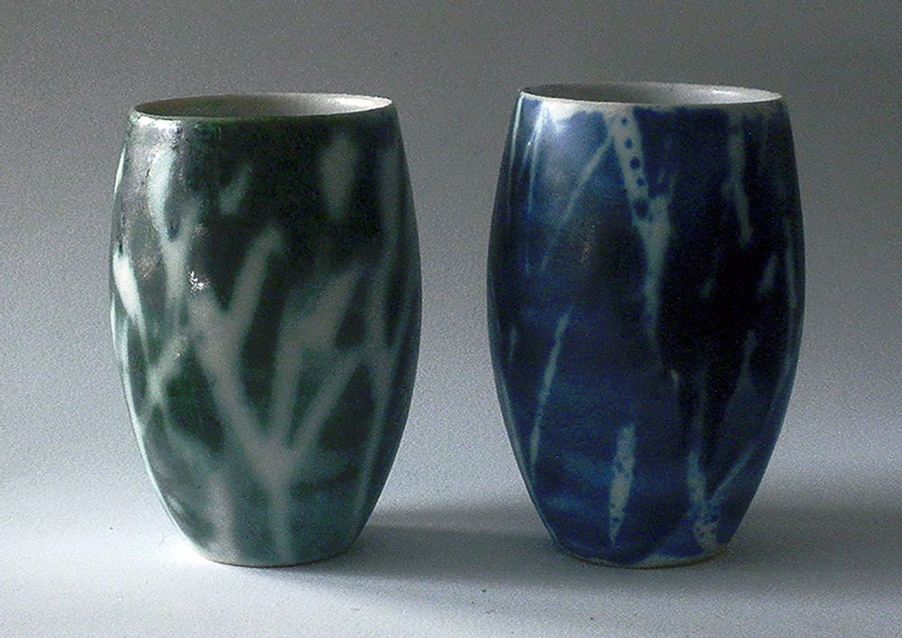 Two slip-cast Tumbler are simple forms of gently curved ceramic drinking vessels glazed in white and then brushed with wax branches and brushed again with fluid interactive ceramic decorating color. One tumbler is brushed in green and the other in blue, The interaction between the flux in the decorating colors and the white glaze created a nature-made effect