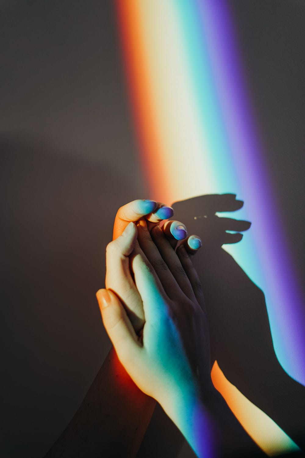 An image of two hands holding each other, with a rainbow light falling across them.
