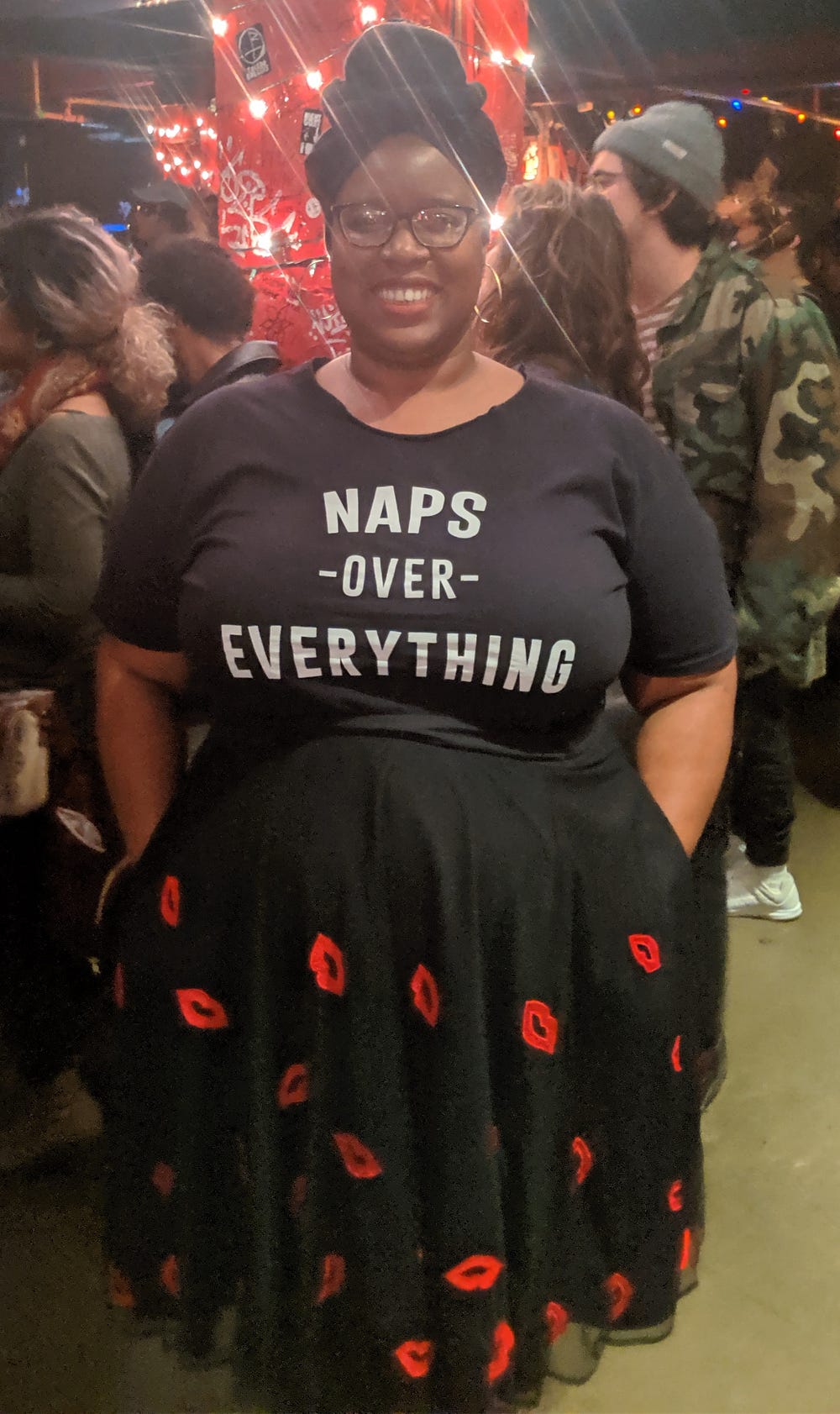 Smiling black woman with black headwrap, tee and skirt. Tee shirt reads: “NAPS OVER EVERYTHING”