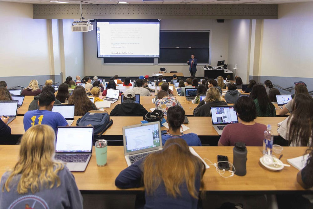 Students fill every seat of a lecture hall. Thomas Garza stands in front of them next to a screen with information on it.