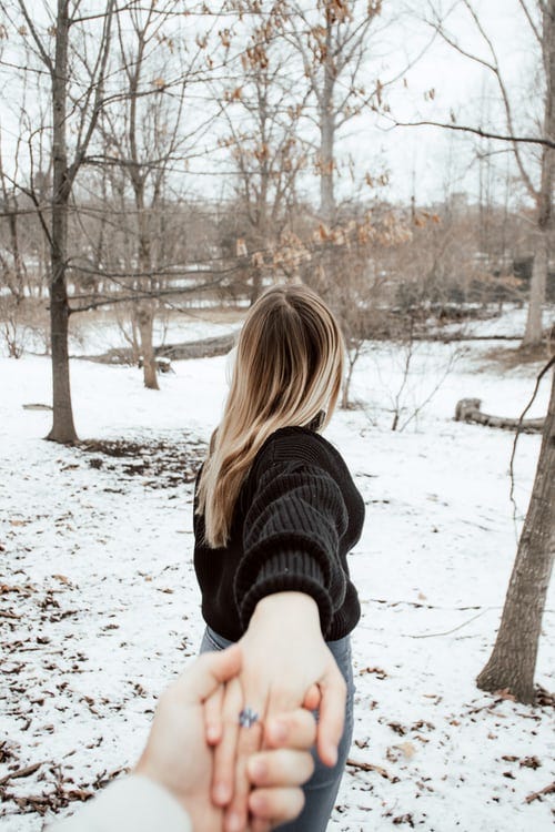 A woman with long hair is holding the hand of someone behind her, and pulling away. They are outside and snow is on the ground.