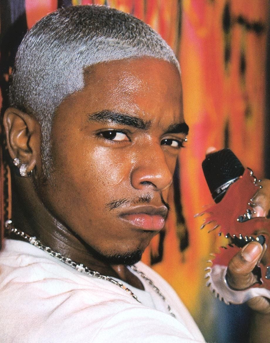 An image of rapper, Sisqó, staring at the camera with a smirk on his face. His hair is short and died white. He has diamond earrings and a silver chain around his neck. He’s holding a microphone in one hand.