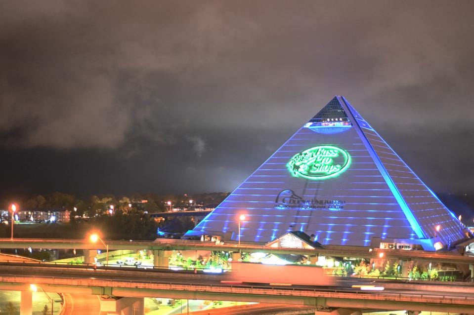 The blue-lit ‘Memphis Pyramid’ towering above the skyline at night, a bright green ‘Bass Pro Shops’ logo is prominent on it.