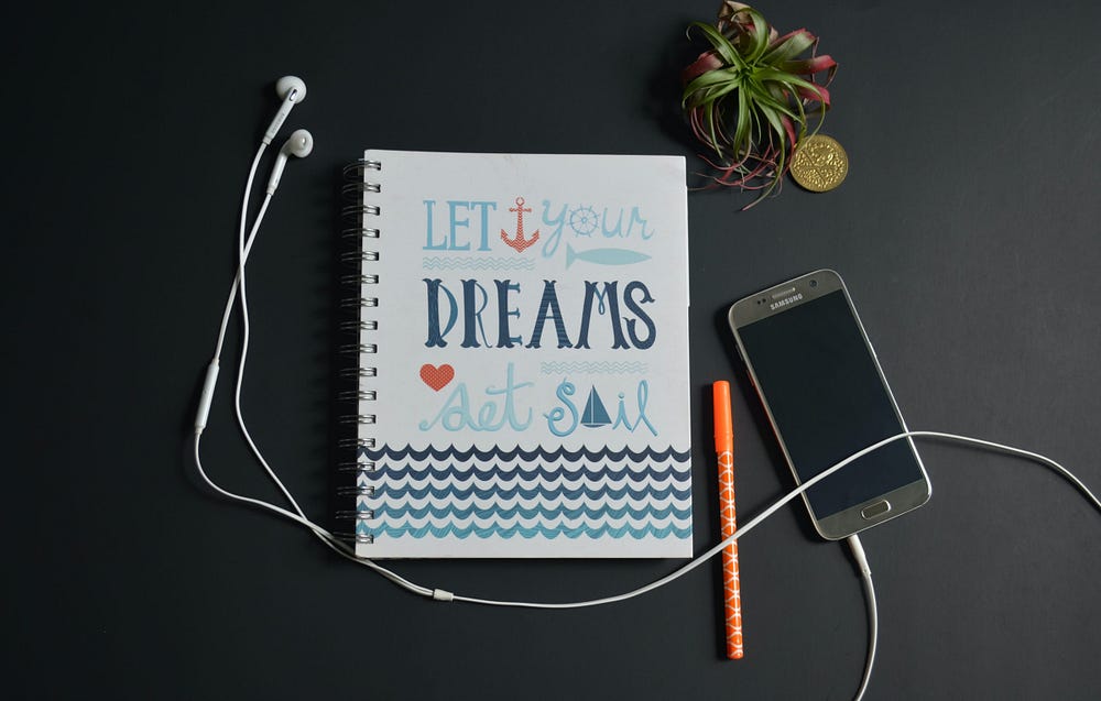A mobile phone with attached headphones accompanied by notebook with title that reads “Let Your Dreams Set Sail.”