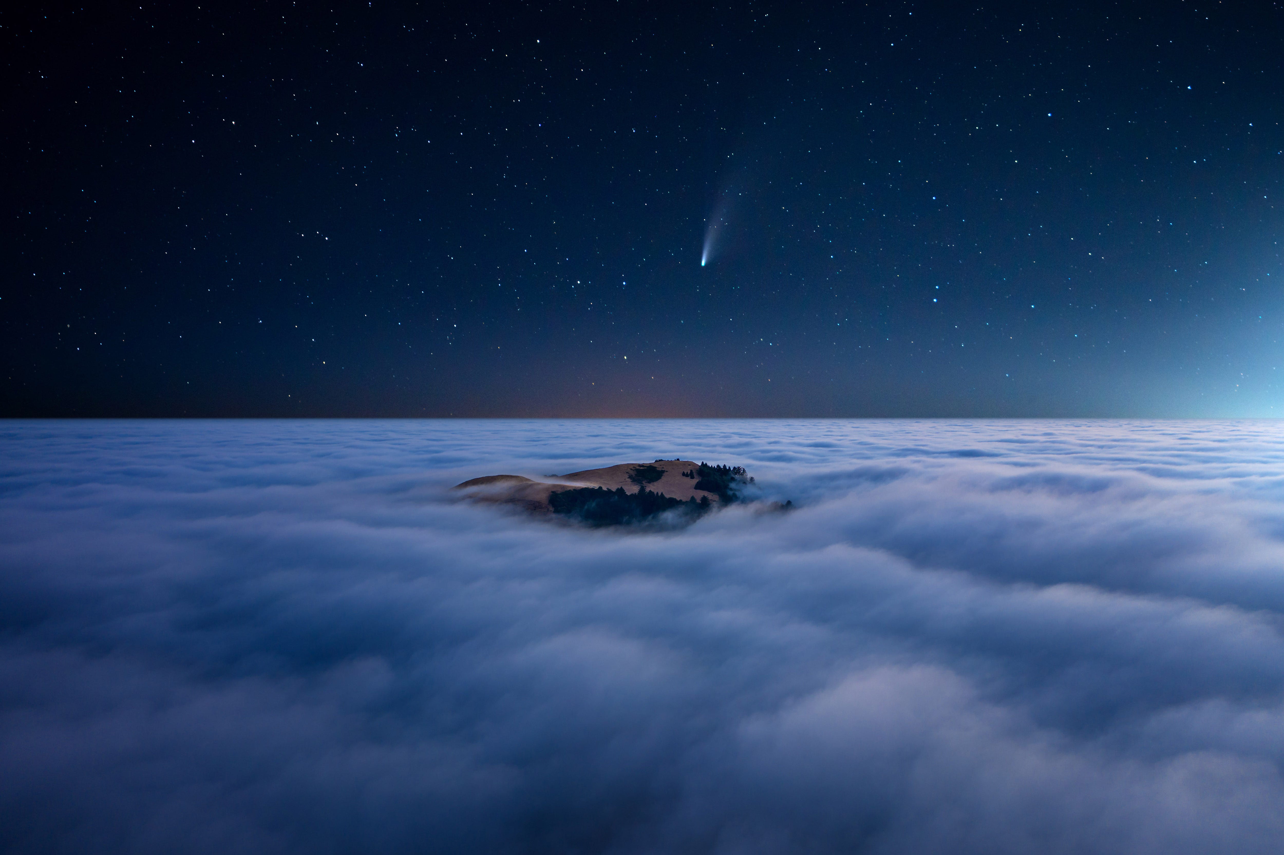 A comet or shooting star appears clearly in the sky above the clouds, which are broken in one spot showing beautiful earth