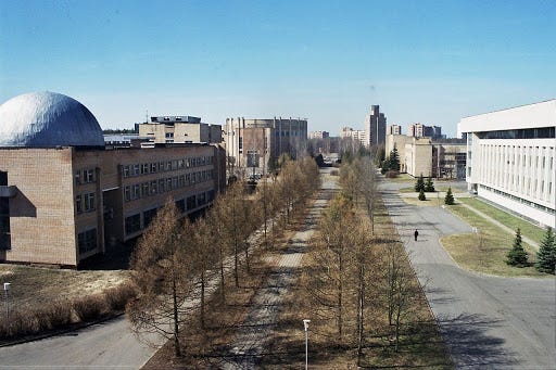 A sidewalk surrounded by several large gray buildings. The closest has a large dome on top