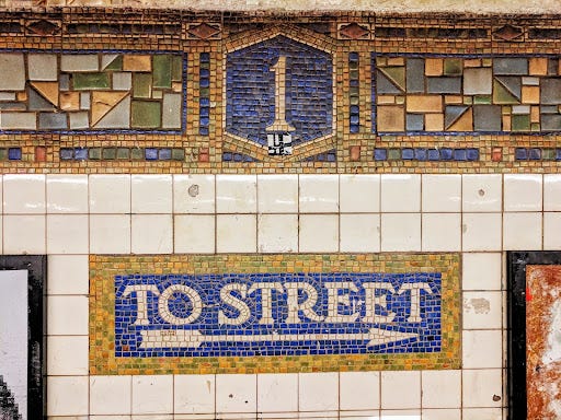 Picture taken of a colorful street mosaic sign in the New York city subway. The mosaic sign tiles are colored yellow and green making up a pattern around the number one and beneath that number one is a rectangle as a sign in the same colored mosaic tiles with a blue backdrop that says “To Street” and has an arrow pointing towards the right.