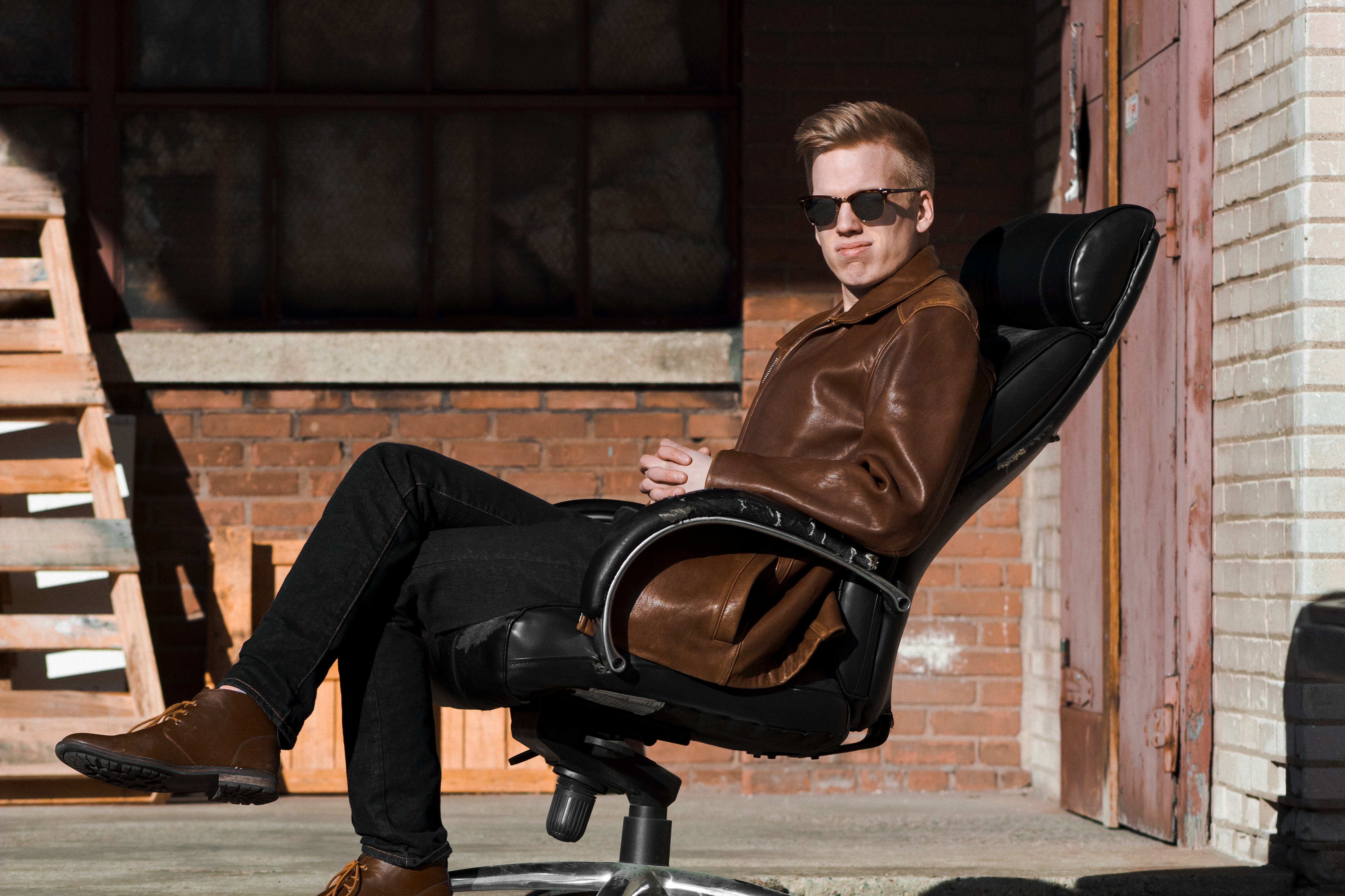 Person wearing brown leather jacket and sunglasses, sitting on black office chair outside