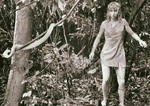 A disheveled girl stands in the jungle in a tattered dress facing a snake on a tree in front of her