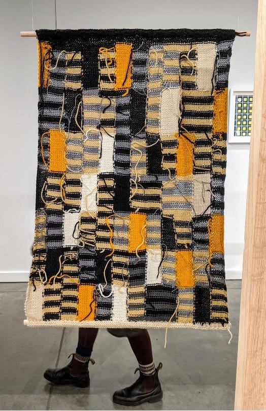 Orange, blue, and black striped patches knit together in a tapestry, which is being held up by someone behind it who is wearing black boots.