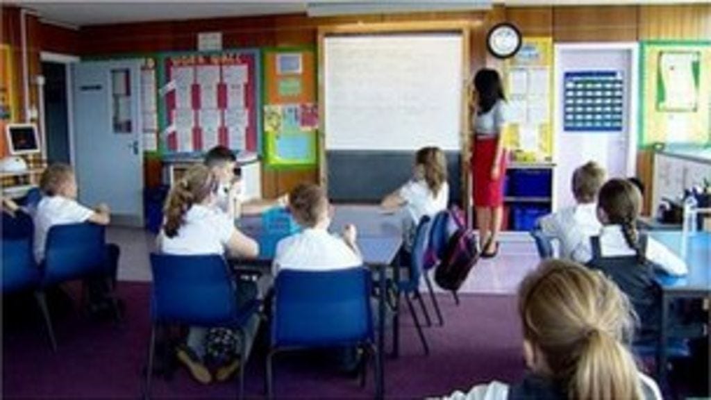 Primary School Children sitting in blue chairs looking at the board and their teacher