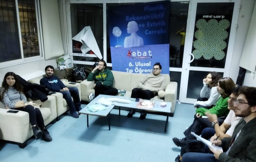 Photo of a student lounge with 8 students sitting in a semi-circle of couches and chairs, with a table between them. A poster with the school name is behind them.