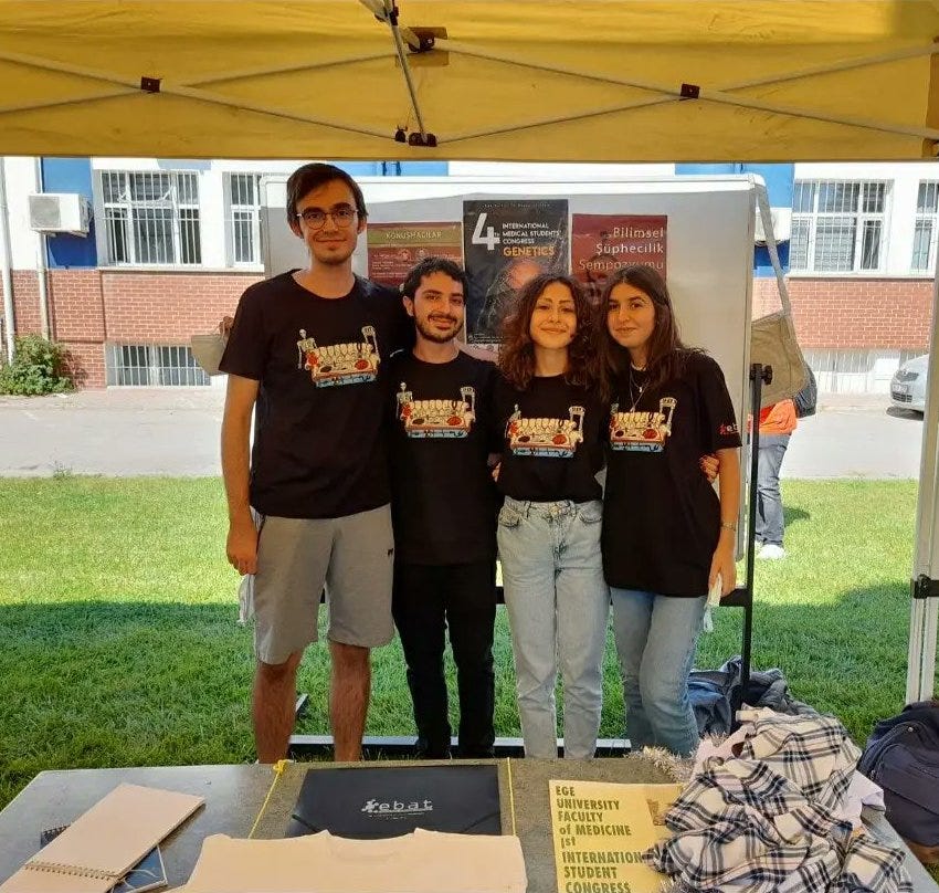 Photo of 4 medical students standing outside a school building underneath a tent, with informational posters behind them and flyers on the table in front of them.