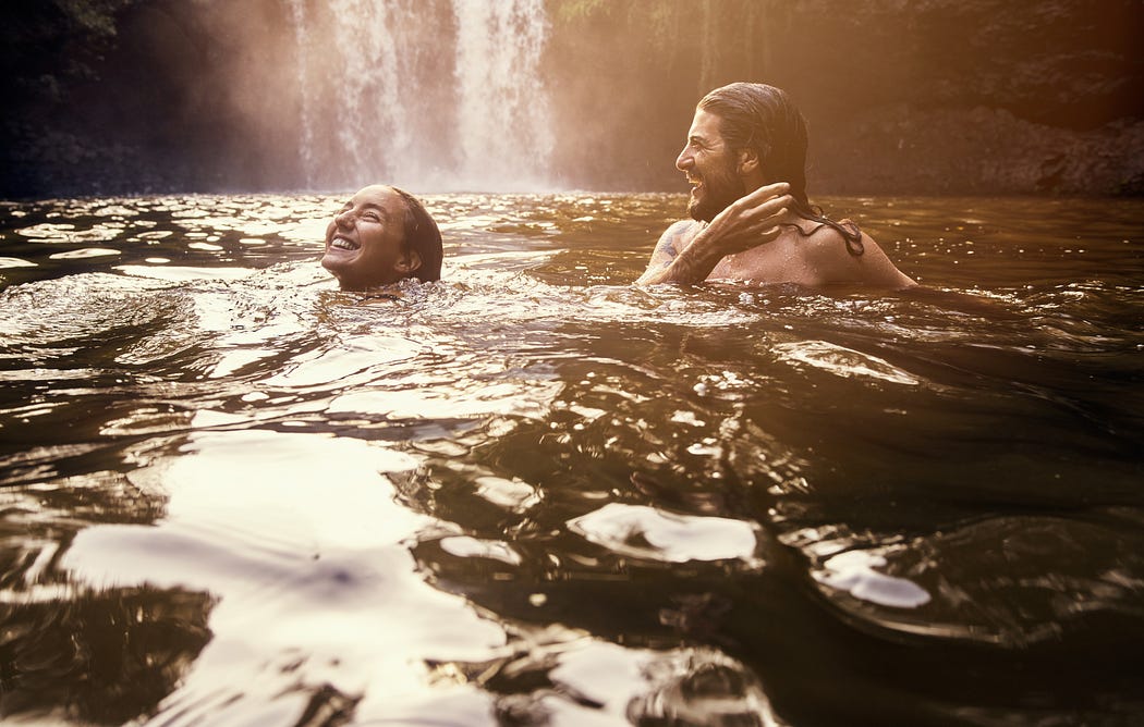 Couple bathing in a scene body of water with a waterfall behind them.