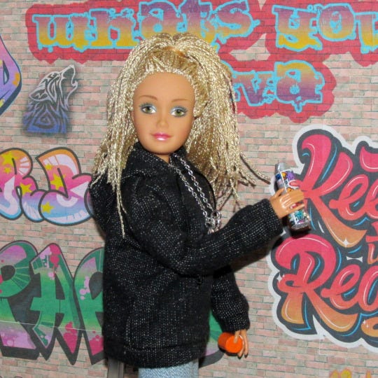 A white female Flavas doll spray-paints a brick wall with graffiti lettering. She’s wearing a fuzzy black coat, makeup, and blonde microbraids.