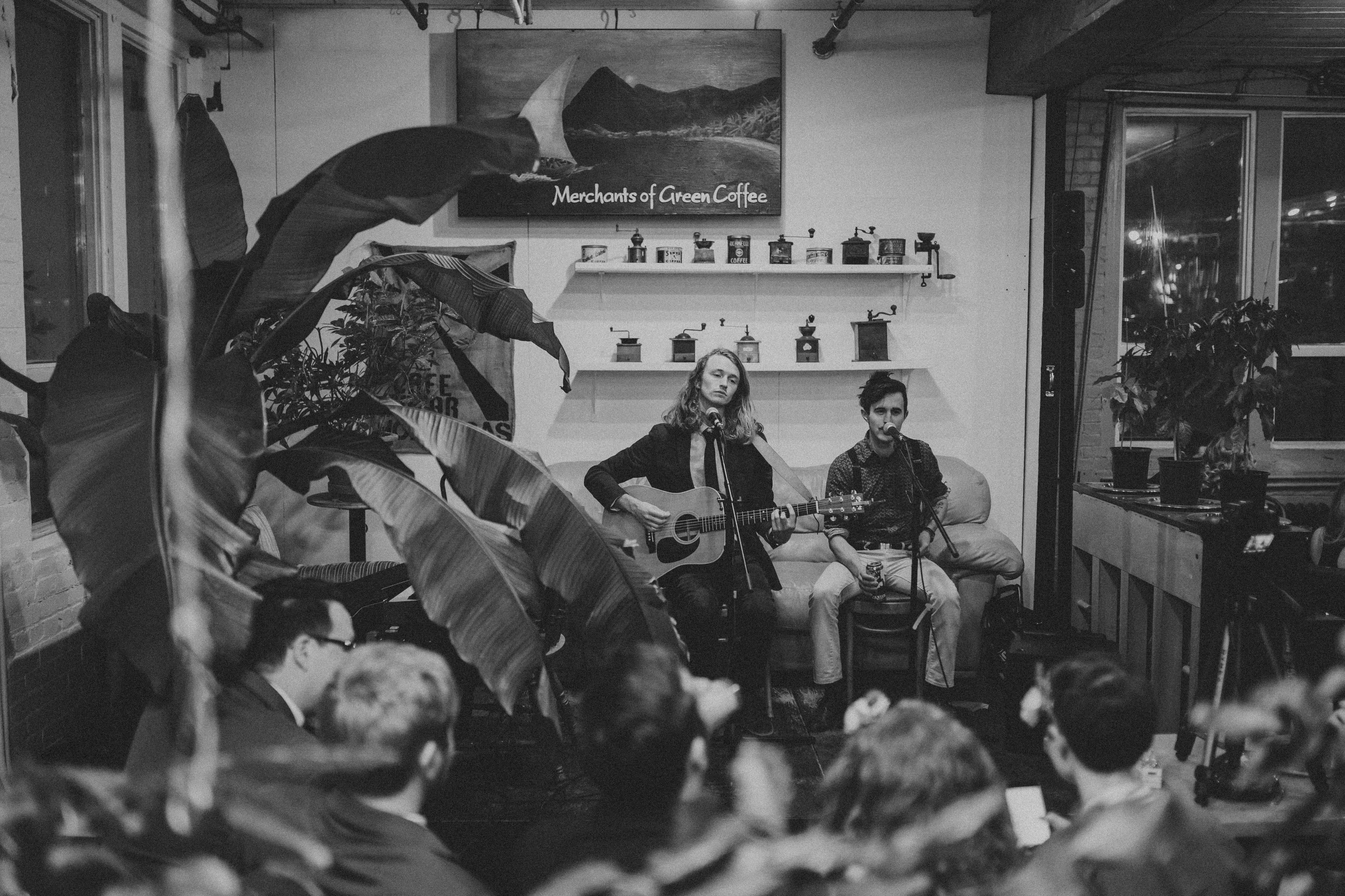 one man sitting on a couch, playing a guitar, singing. Beside him is another man singing. People are sitting opposite of them.