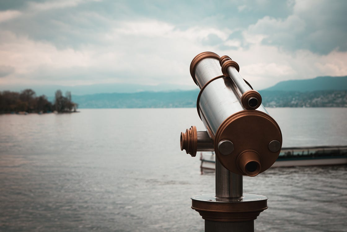 telescope pointed into the distance across a large lake against a gray sky with mountains on the horizon