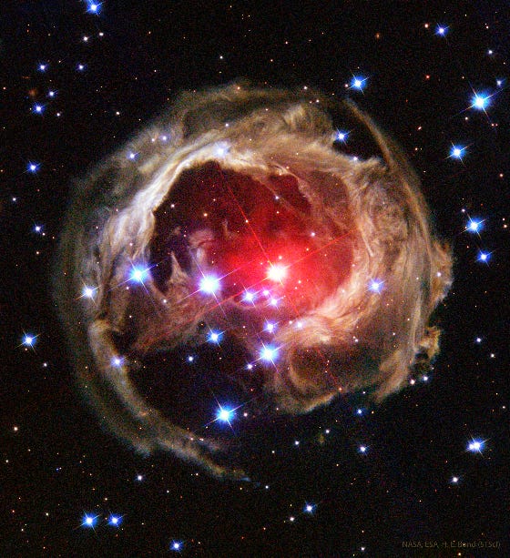 A red star with a red halo sits near the center of a stellar landscape. It creates a point on an arrow-like grouping of blue-white stars from the left corner. Around the red star is an incomplete “orb” of tan dust and stellar material, wisps circling around the red star with gaps particularly in the bottom and in the center. Other stars like in the background.