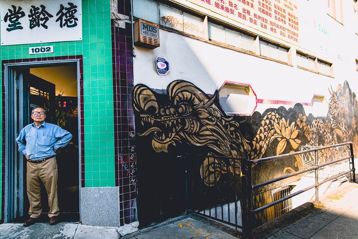 A photo of an Asian man standing in front of a store in Oakland Chinatown. There is a long golden and black dragon mural to the right of him. He is standing in front of a green wall, below a sign written in Chinese.