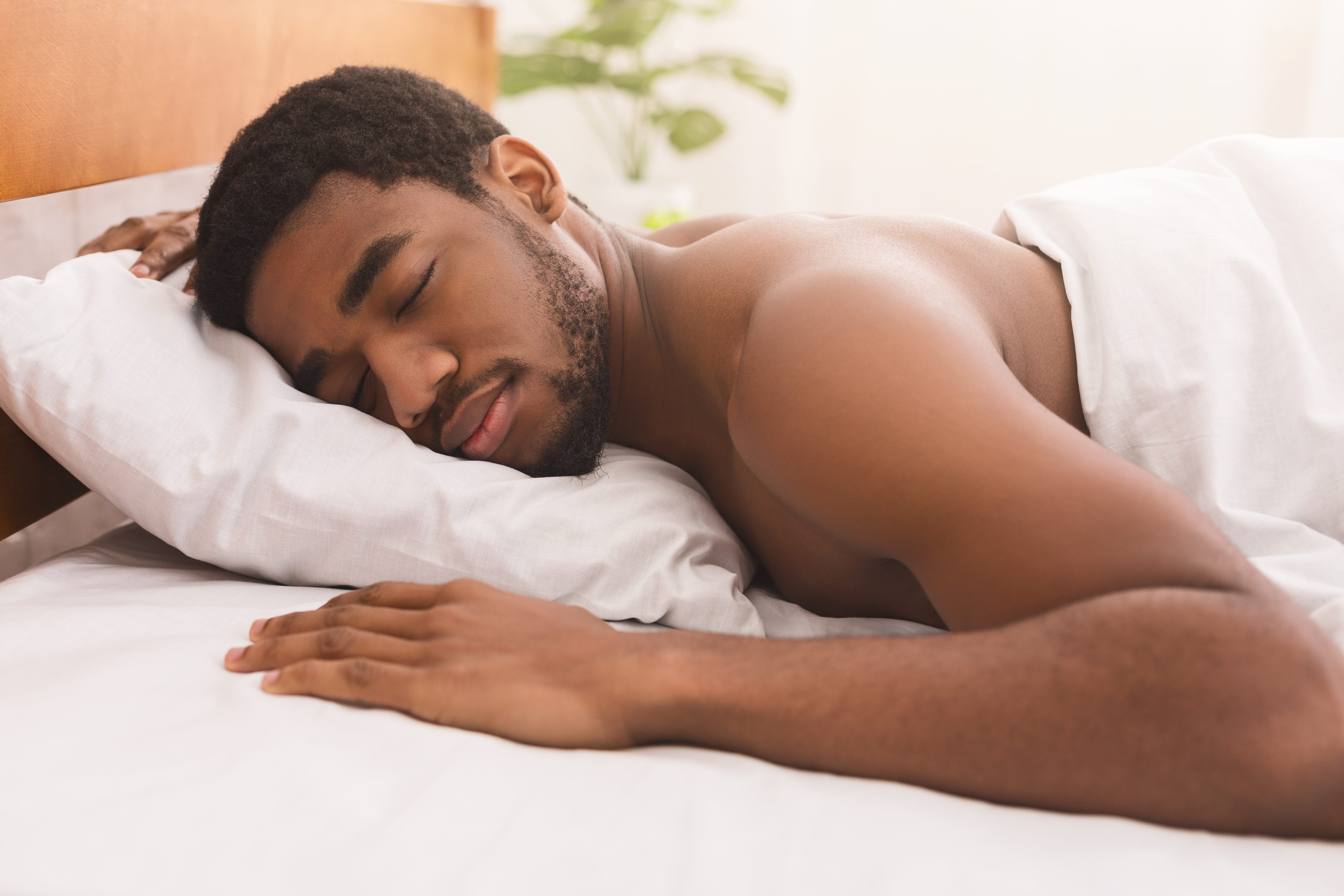 An African-American man sleeping naked under the covers on his stomach in bed at home.