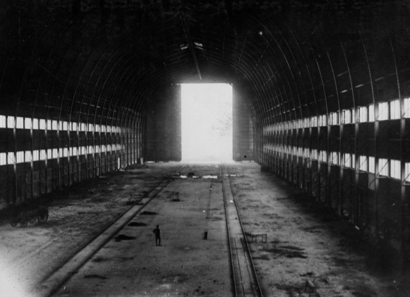 A black and white photo showing an internal view of a Zeppelin shed looking from the cavernous interior towards the sliding doors through which daylight shines. A small figure is standing on the ground, dwarfed by the structure around him.