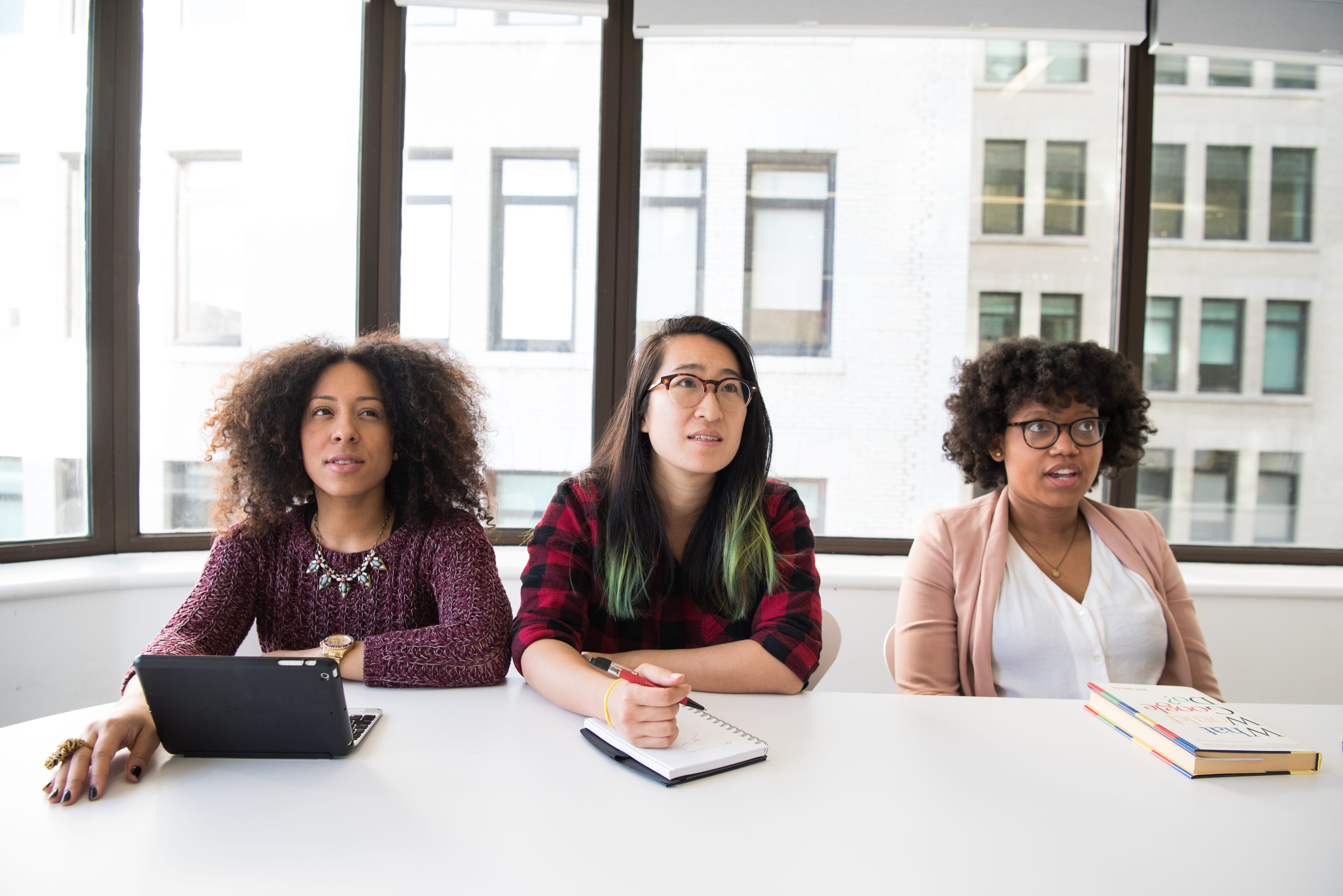 Three woman of color sitting at a table in a windowed conference room, looking surprised and concerned.