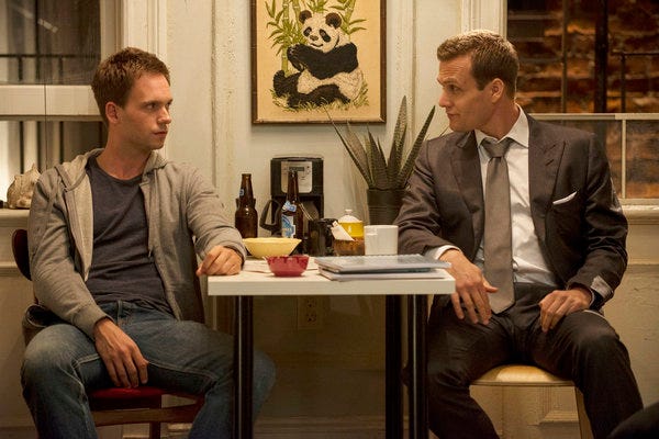 Mike and Harvey sitting at a table in Mike’s apartment, Mike is wearing a t-shirt and hoodie, and Harvey is wearing a suit with the tie loose. They both look stoned