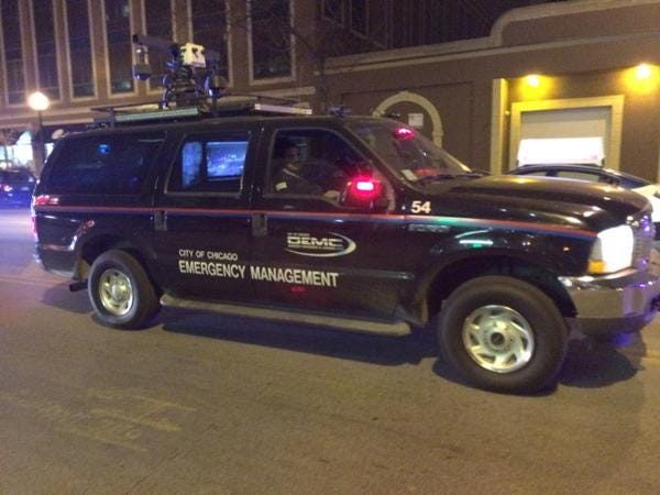 A black truck with a red stripe and the text ‘Emergency Management’ on the side. It has red glowing police lights on the side view mirrors. On the roof is a strange black antennae apparatus.