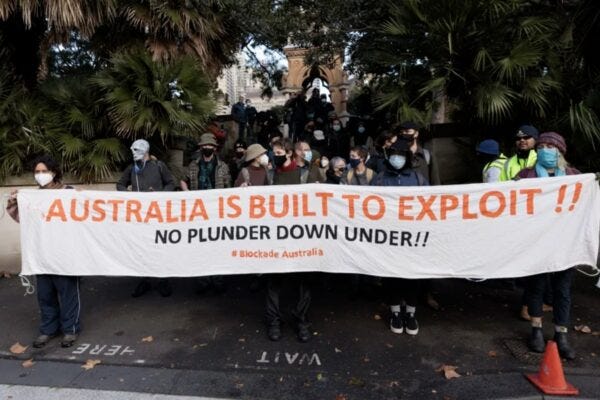 Crowd of people holding banner reading Australia is built to exploit!! No plunder down under #BlockadeAustralia