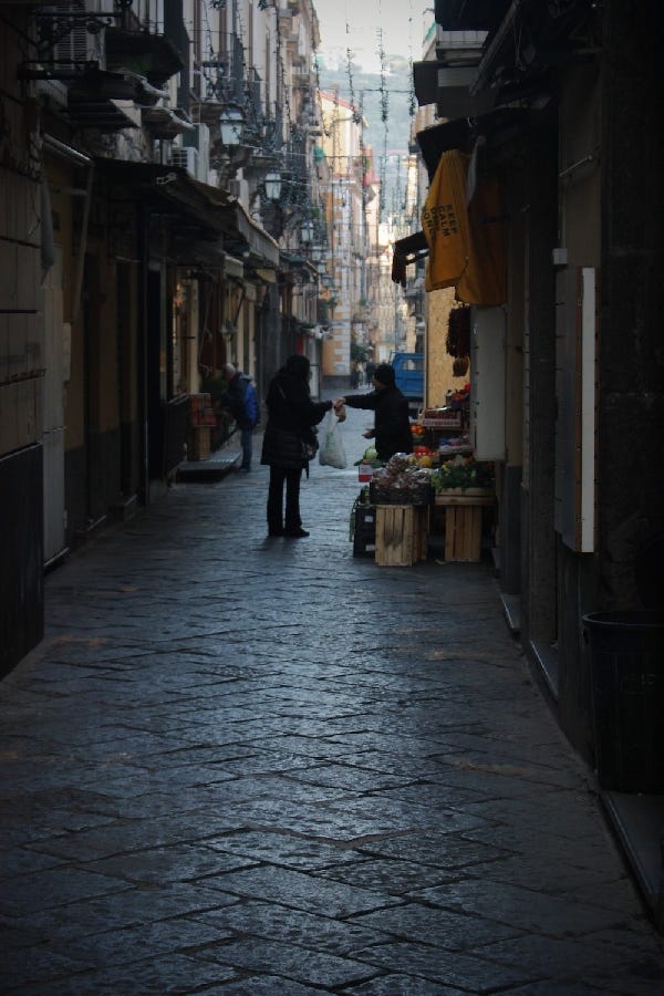 A shop owner hands a customer some produce in a narrow alley at dawn