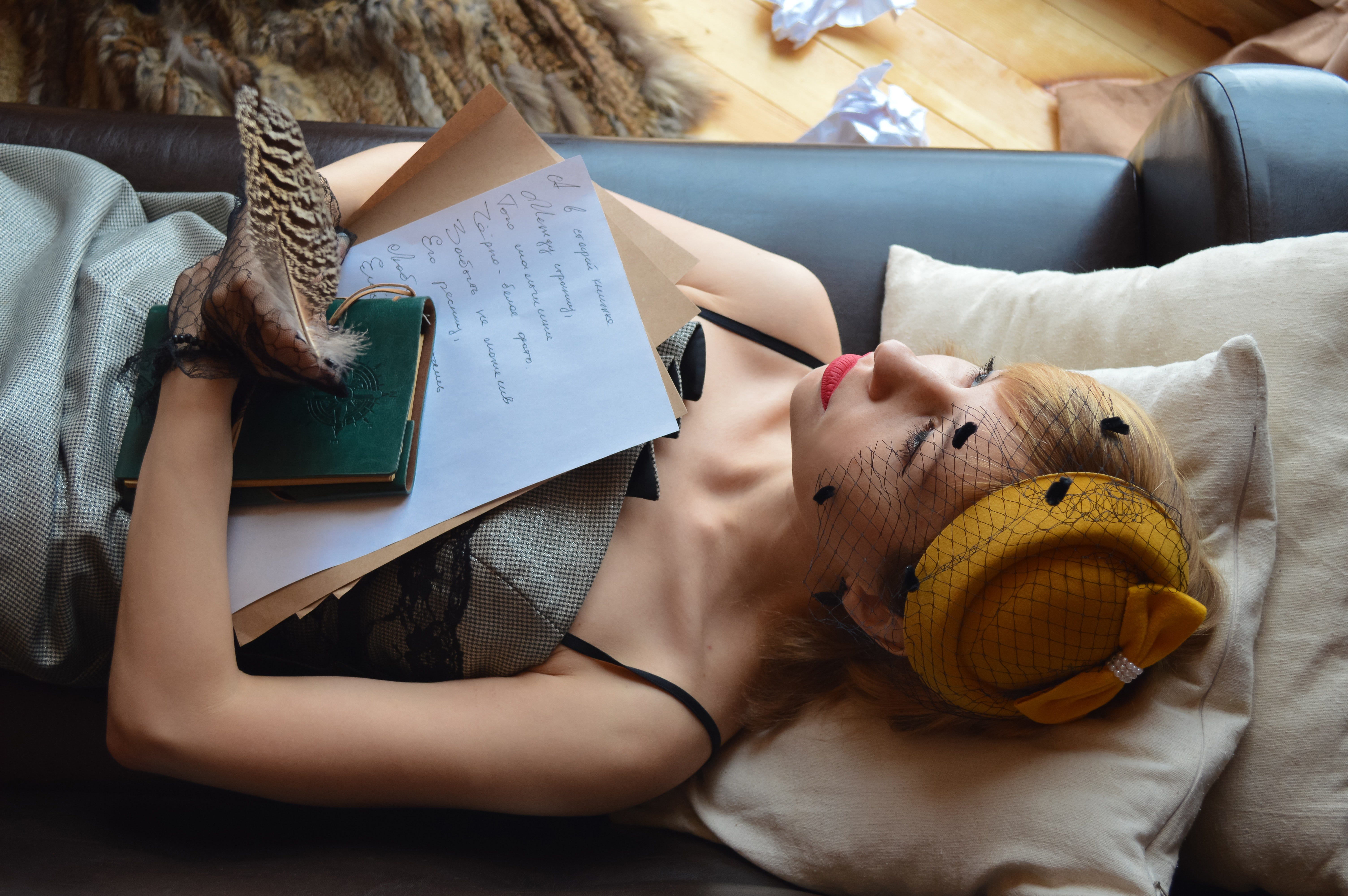 Elegant woman dressed in vintage attire, laying on a couch. She is holding papers, a notebook, and a feathered pen. Crumpled papers lay on the wooden floor behind the couch.