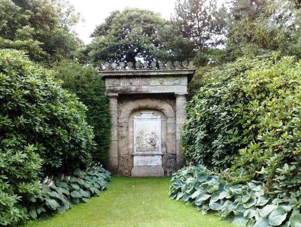 A grassy path between lush shrubbery and trees leads to a stone sculpture with ornate carvings and inscriptions.