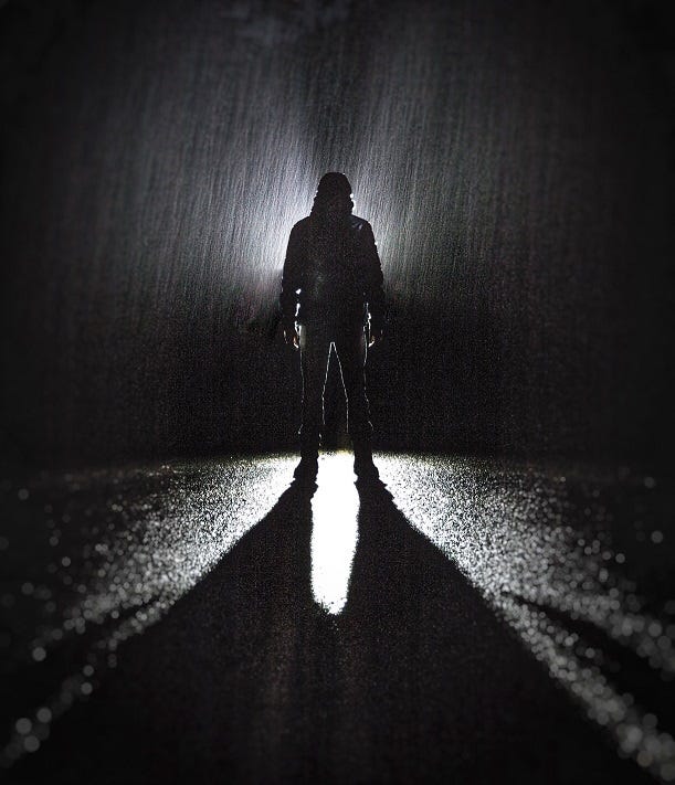 A black and white photo of a siloette of a person standing in the middle of a rainy road with headlights illuminating