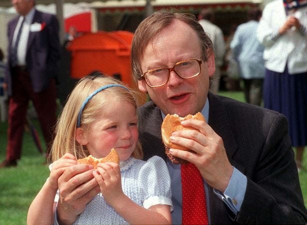 Colour photo taken outdoors on a sunny day of a four-year-old blond white girl eating a beef burger in a white bread bun next to a middle aged white man with light brown hair and glasses (John Gummer) also eating a beef burger in a white bread bun.