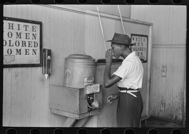 Black man drinking from a water fountain labeled ‘Coloured’ in between signs for “White women, colored women’ and “White men, colored men’