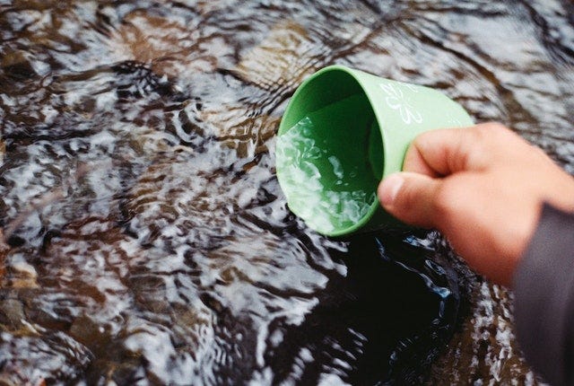 A hand is pictured holding a green mug that is being dipped into a stream of water.